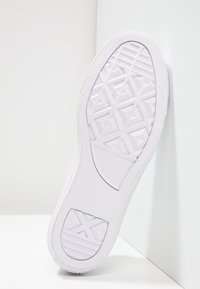 Semelle de basket blanche Converse All Star avec un motif géométrique distinctif, contre un mur et un sol blancs en angle.