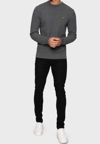 Suéter gris de punto con cuello redondo y un pequeño logotipo amarillo en el pecho, combinado con pantalones ajustados negros y zapatillas deportivas blancas.