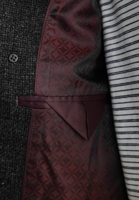 Forro de chaqueta de tela texturizada gris con un interior burdeos y estampado, y un bolsillo lateral, combinado con una camisa de rayas debajo.