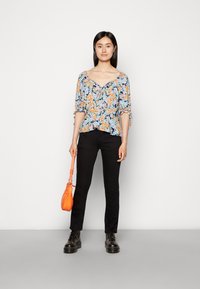 Blusa floral com mangas bufantes, padrão azul, laranja e branco; combinada com calças pretas e botas pretas com atacadores; segurando uma mala laranja.