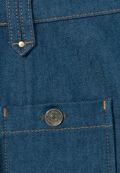 Tejido de mezclilla en azul oscuro con textura visible. Presenta un botón a presión plateado y un hilo en contraste color marrón claro cerca de un bolsillo pequeño.