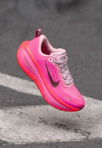 Ljusrosa Nike ZoomX-löparsko med grå swoosh, svävande över asfalt med vit vägmarkering.