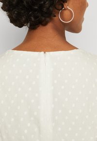 Λευκό φόρεμα με υφασμάτινη πουά υφή, διαθέτει φερμουάρ στην πλάτη και στρογγυλή λαιμόκοψη. Το ύφασμα φαίνεται ελαφρύ και λείο.