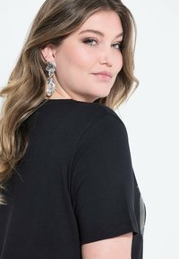 Svart t-shirt med korta ärmar, med ett strukturerat mönster på baksidan. Modellen har långt, vågigt hår och bär stora, metalliska örhängen.