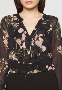 Svart blommig blus med volangkrage, prydd med rosa blommor och gröna blad. Genomskinliga långa ärmar och knappdetalj framtill.