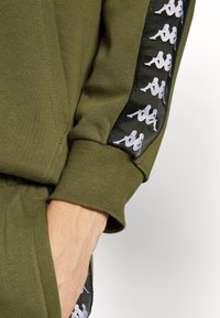 Arm met olijfgroene sweatshirt met geribbelde manchetten en een zwarte streep met herhaalde witte Kappa-logo's langs de mouw.