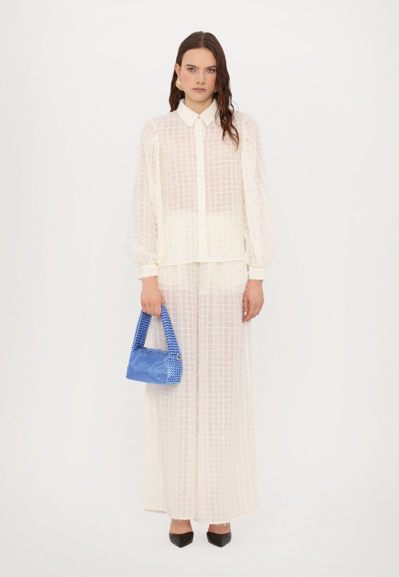 Blusa bianca con texture e motivo a griglia, abbinata a una gonna coordinata e a una borsa blu. Indossata con tacchi a punta neri.