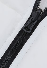 Witte doorgestikte stof met een zwarte rits. De rits heeft een getextureerd trekkoord en merklogo, met contrasterende stiksels langs de naden.