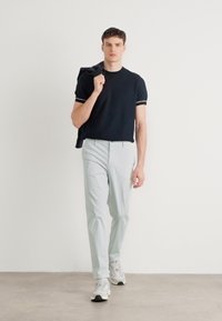 Tricou negru cu margini albe, pantaloni gri deschis și adidași gri. Modelul ține o jachetă neagră pe umăr. Design simplu, modern.