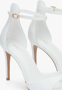 Witte hoge hakken sandalen hebben een gladsynthetisch uiterlijk, een open teenontwerp en een slanke enkelriem met een gouden gesp.