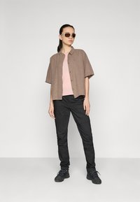 Bruine kortblijvende shirt, roze T-shirt, zwarte cargobroek, zwarte sneakers, oversized beige zonnebril, gladde stof, relaxte pasvorm.
