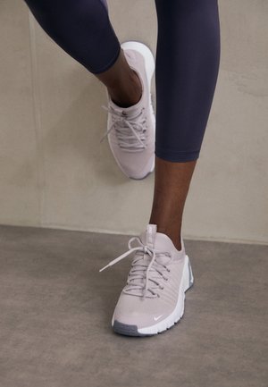 Light grey sport shoes with a textured upper, lace closure, and white sole, paired with dark leggings. Minimalist design and smooth accents.