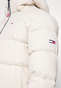 Chaqueta de plumas acolchada beige con capucha, cierre de cremallera y pequeños parches de logotipo en rojo, blanco y azul marino en el pecho y la manga izquierda.