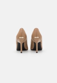 Un paio di scarpe con tacco alto beige con sottili tacchi a stiletto viste da dietro su uno sfondo bianco.
