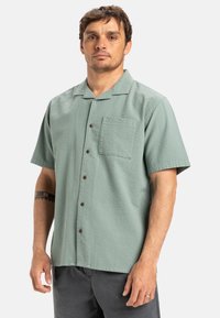 Short-sleeve, textured button-up shirt in light green with a chest pocket and brown buttons, featuring a relaxed collar design.