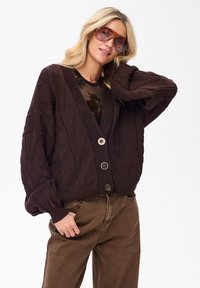 Brązowy sweter z dzianiny z dekoltem w kształcie litery V, ozdobiony diamentowym wzorem, z prążkowanymi brzegami i dwoma dużymi guzikami. Połączony z brązowymi spodniami.