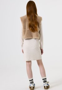 Jonge vrouw met lang rood haar draagt een beige jurk, een tan bontvest, witte sokken met zwarte tekst en sneakers met panterprint, staand tegen een witte achtergrond.