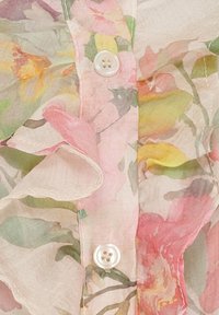 Blommig blus med volantkanter, genomskinligt tyg i pastellfärger, med en vit knapp i mitten. Subtila texturer och mönster.