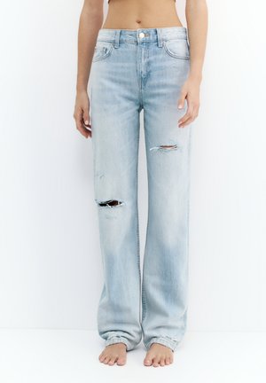 Frau trägt hellblaue, hoch taillierte, zerrissene Jeans, steht barfuß vor weißem Hintergrund, mit freiem Bauch.