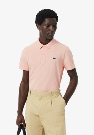 Polo de color rosa claro con cuello clásico, tapeta de tres botones y un pequeño logo azul marino en el pecho, combinado con pantalones beige.