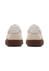 Baskets en cuir suede blanc avec une semelle en caoutchouc brun, présentant un design arrondi et un logo en relief sur le talon. Texture douce, style minimaliste.