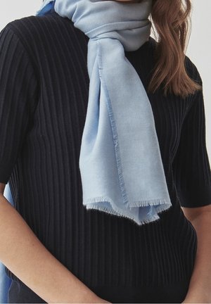 Femme portant une écharpe bleu clair à franges nouée autour du cou, par-dessus un haut noir côtelé à manches courtes, raccourci à la taille.