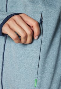 Casaco azul claro com fecho éclair, com tecido texturizado, acabamento em azul marinho e um bolso lateral com um rótulo verde perto da bainha.