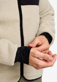 Fleecejacka i krämfärg med en svart nylonbröstficka. Har stretchiga svarta muddarmar och dragkedja fram. Mjuk, strukturerad tyg.