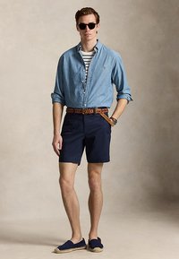 Férfi napszemüvegben, farmer ing csíkos póló fölött, tengerészkék rövidnadrág fonott övvel, espadrille cipőben, egy kézzel a zsebében áll.