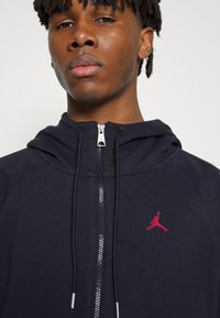Schwarze Zip-Hoodie mit Kapuze, silbernem Reißverschluss und rotem Jumpman-Logo auf der Brust. Glatter Stoff mit fitted Design.