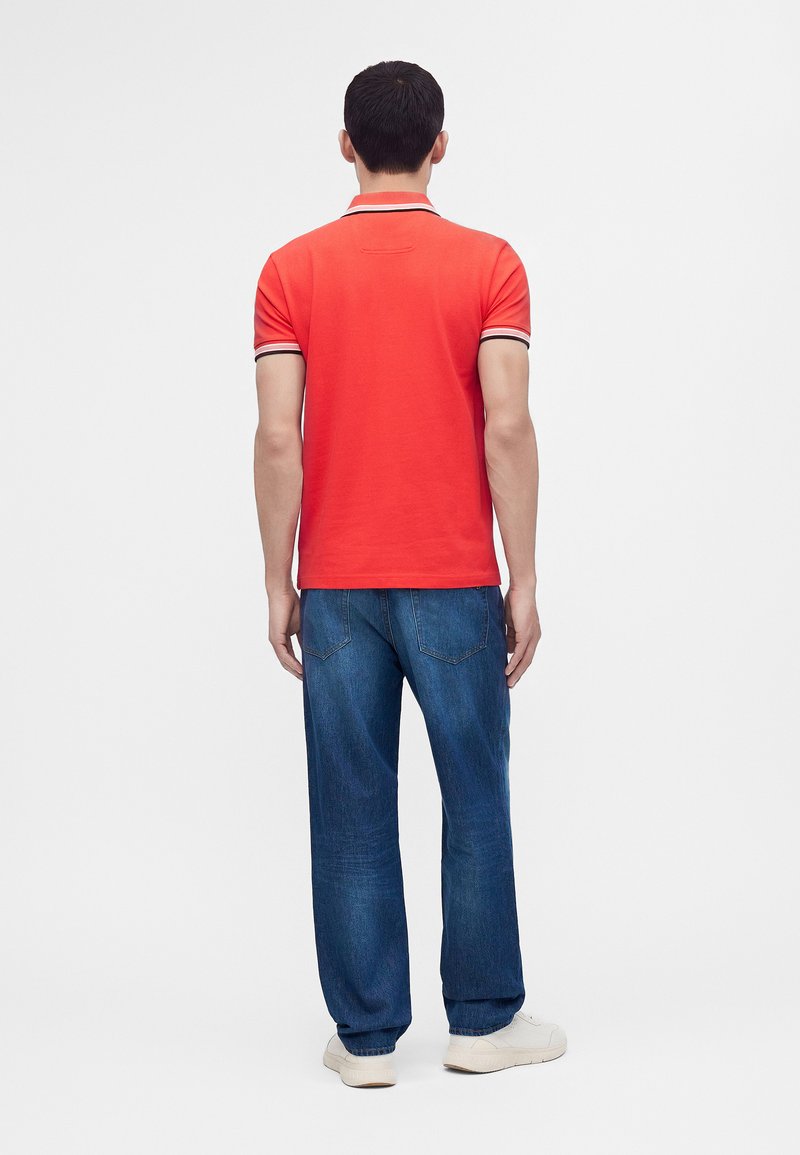 Homme portant un polo rouge à col rayé et un jean bleu, se tenant de dos sur un fond blanc uni.