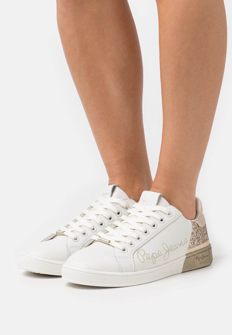 Baskets blanches avec une tige en cuir lisse, des accents métalliques et un panneau de talon en paillettes dorées texturées. Présente une fermeture à lacets.