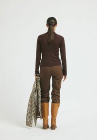 Smeđa longsleeve majica u kombinaciji s smešnim hlačama i čizmama do koljena u boji tamne bež. Drži uzorčani bež i smeđi šal od zmijske kože.