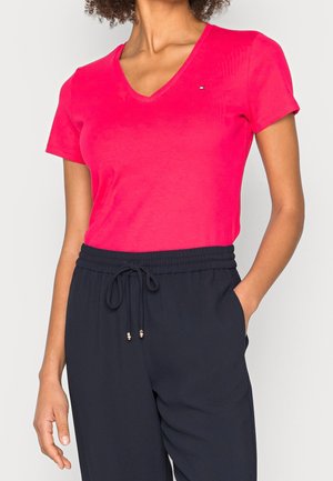 Vrouw draagt een felroze T-shirt met korte mouwen en V-hals en een zwarte broek met trekkoord, staand met één hand in de zak tegen een lichte achtergrond.