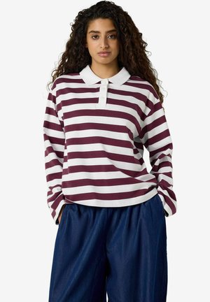 Femme aux cheveux bouclés portant un polo à manches longues rayé blanc et bordeaux et un pantalon ample bleu foncé, mains dans les poches.