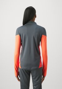 Haut de sport à manches longues avec un haut gris et des manches orange vif, fabriqué en tissu lisse et flexible, doté d'un col et d'une coupe ajustée.