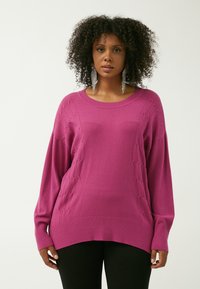 Übergröße magentafarbener Pullover mit geripptem Saum und Bündchen, der ein strukturiertes Muster an den Ärmeln und leichtes Strickmaterial aufweist.