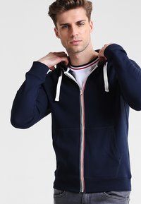 Sudadera con cremallera en color azul marino, con cordones blancos, rayas rojas y blancas en contraste a lo largo de la cremallera y bolsillos frontales. Textura suave.