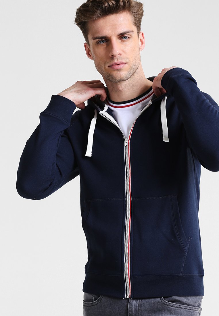 Sudadera con cremallera en color azul marino, con cordones blancos, rayas rojas y blancas en contraste a lo largo de la cremallera y bolsillos frontales. Textura suave.