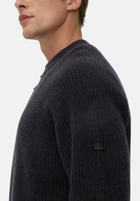 Dunkelgrauer, gerippter Pullover mit rundem Ausschnitt und einem dezenten Logo-Patch am linken Ärmel. Weiche Textur und figurbetonte Passform.