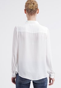 Blusa blanca de manga larga con una textura suave, que presenta un panel en el canesú, un dobladillo curvado y puños abotonados. Combinada con jeans azules.
