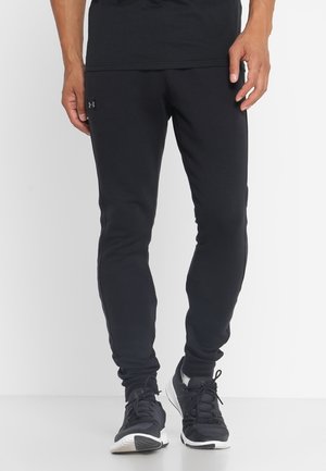 Mann trägt schwarze schmal geschnittene Jogginghosen und schwarze Sportschuhe, steht vor weißem Hintergrund.