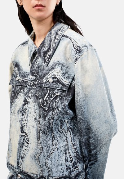 Denim-takki, joka on vaalean sininen mustilla abstrakteilla kuvioilla, klassisella kauluksella ja kahdella etutaskulla, joissa on hopeanväriset napit.