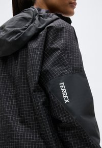 Personne portant une veste d'extérieur noire à motif quadrillé avec capuche, affichant le logo "TERREX" sur la manche, sur un fond uni.