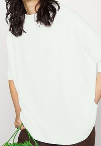 Luźna biała bluzka z wysokim dekoltem, krótkimi rękawami i bocznymi kieszeniami. Teksturowany materiał układa się płynnie, uzupełniony zieloną torbą.