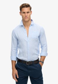Camisa de lino azul claro, con botones, bolsillo en el pecho, mangas enrolladas y detalle de puños, combinada con pantalones azul oscuro y un reloj.