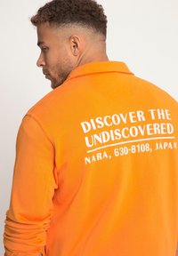 Camisa de manga larga naranja con cuello polo. Presenta texto blanco en la parte de atrás: "DESCUBRE LO NO DESCUBIERTO, NARA, 630-8108, JAPÓN." Textura suave.