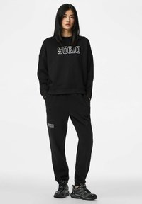 Jauna moteris stovi su rankomis kišenėse, dėvi juodą džemperį ir laisvalaikio kelnes su užrašu „SOHO“ bei juodus sportinius batelius.