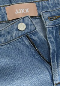 Jeans de mezclilla azul claro con cremallera, botón plateado y costuras naranjas en contraste. La etiqueta dice "JJXX" sobre un fondo rosa claro.