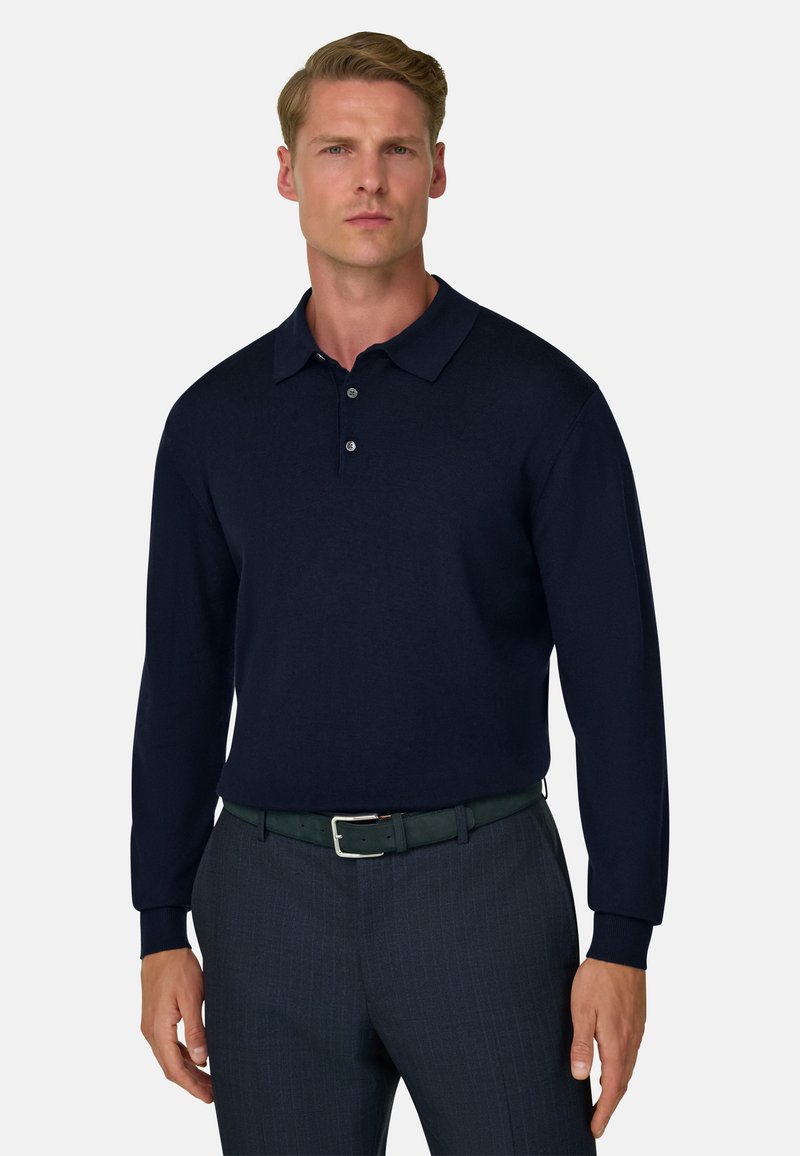 Homme portant un polo manches longues bleu marine foncé rentré dans un pantalon gris avec une ceinture noire, debout devant un fond clair uni.